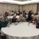 Tables of members are attempting an exercise with sticks, while the table in the foreground has four women laughing and smiling as they try